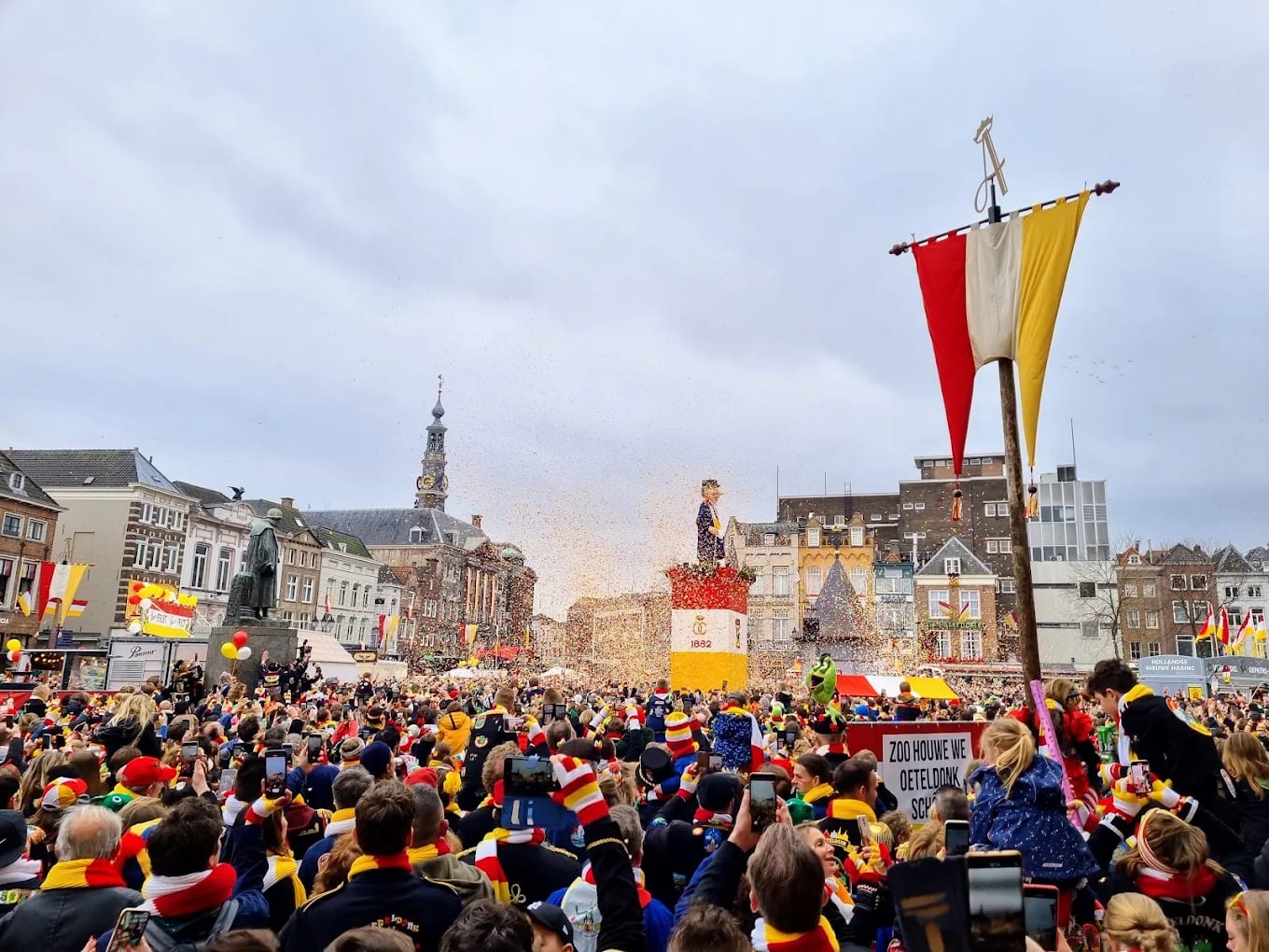 Volle Markt in Oeteldonk meej rood-wit-gele vlaggen en Knillis op z'n sokkel